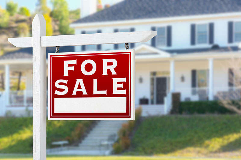 A red and white For Sale sign is posted in front of a blurred, large two-story suburban house with white siding, black shutters, and a front porch.
