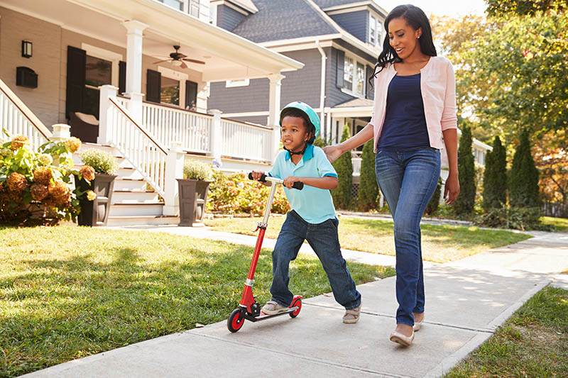A young boy wearing a helmet rides a red scooter on a sidewalk, while an adult woman walks beside him, gently holding his arm. They are in a residential neighborhood with houses and greenery in the background.