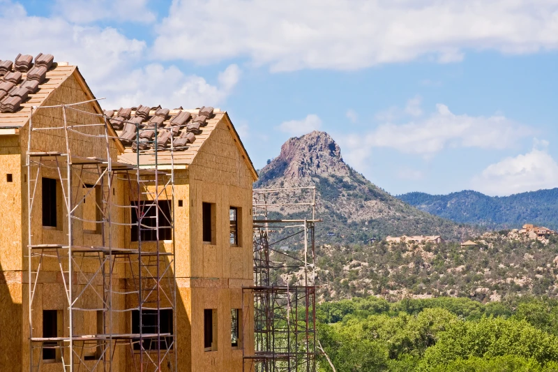 A wooden house under construction with scaffolding stands in the foreground, while green trees and a rocky mountain peak are visible in the background under a partly cloudy sky.